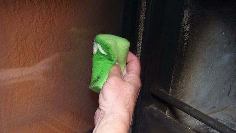 Eine Hand hält einen grünen Schwamm, der gerade ein Ofenfenster mit Scheuermilch reinigt, in einem hellen Raum.