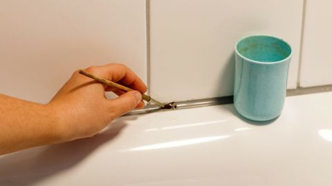 Bringe Deine Acryl- und Silikonfugen zum Glänzen: Mische Silikonöl mit Wasser und trage die Lösung mit einem Pinsel auf. Der Artikel zeigt, wie einfach es geht und die Fugen wieder wie neu erstrahlen. Hand pinselt Acryl/Silikonfugen in Bad; blauer Becher daneben dient als Behälter.