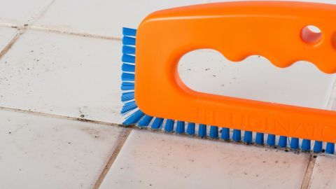 Ein orangefarbener Fugenreiniger wird über schmutzige Fliesenfugen gezogen, um effektiv zu reinigen und den Dreck zu entfernen.