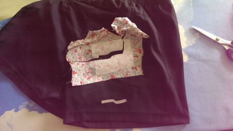 Ein schwarzes Kleidungsstück liegt auf einem Tisch. Vliesofix wird auf die vorgeschnittenen Teile aufgebracht und aufgebügelt.