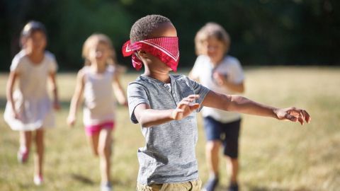 Ein Kind mit einem roten Tuch vor den Augen bewegt sich auf einer Wiese, während andere Kinder hinter ihm spielen.
