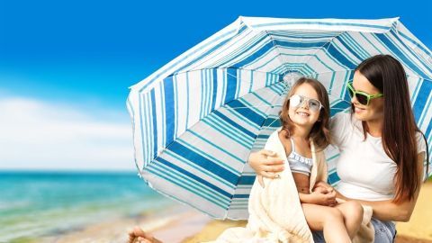 Eine Mutter sitzt mit ihrer Tochter unter einem Sonnenschirm am Strand, um sich vor der Sonne zu schützen.