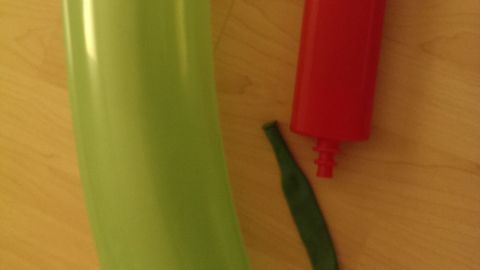 Ein grüner Luftballon in Form einer Gurke liegt neben einer roten, mechanischen Pumpe auf einem hellen Holztisch.