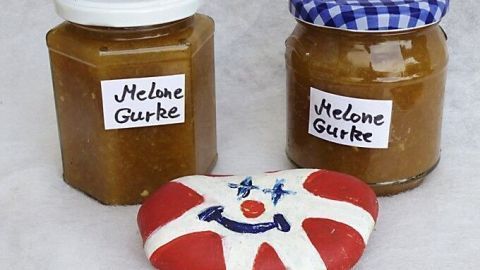 Zwei Gläser Melonen-Gurken-Marmelade stehen dekorativ nebeneinander, während ein bemalter Stein mit einem Clowns-Gesicht die Szene ergänzt.