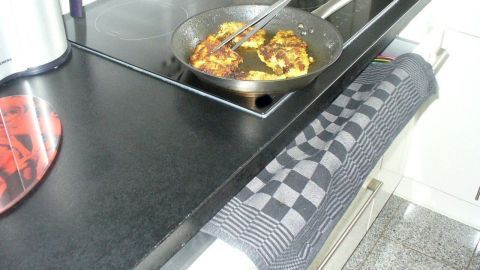 Eine Pfanne mit Braten auf einer modernen Küchenarbeitsfläche, während ein Geschirrtuch bereitliegt, um Spritzer aufzufangen.
