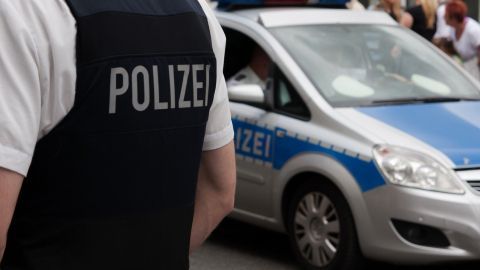 Ein Polizist steht in Uniform neben einem Polizeiwagen, während Menschen im Hintergrund in einer urbanen Umgebung sichtbar sind.