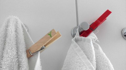 Weiße Handtücher hängen an einem Haken, jeweils mit einer Wäscheklammer befestigt, auf einem hellen Hintergrund.