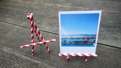 Eine Mini-Staffelei aus Strohhalmen hält ein Bild mit einer Landschaft. Sie steht auf einer rauen Oberfläche.