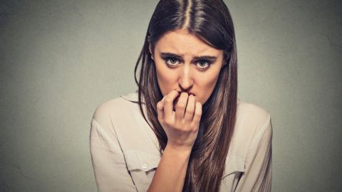 Eine besorgte Frau beißt auf ihre Fingernägel, während sie ängstlich in die Kamera schaut, vor einem neutralen Hintergrund.