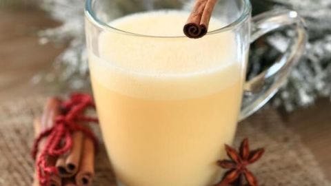 Ein Glas Eierpunsch steht auf einem Jutetuch. Dazu eine Zimtstange und Sternanis, im Hintergrund winterliche Dekoration.
