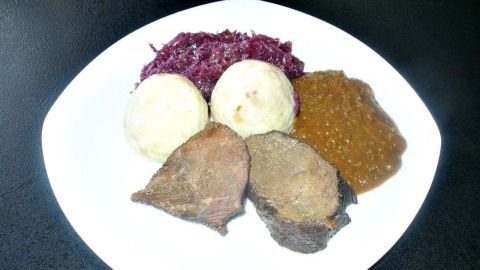 Saftiger Rotweinbraten liegt zusammen mit Knödeln, Rotkohl und Soße auf einem weißen Teller vor schwarzem Hintergrund.