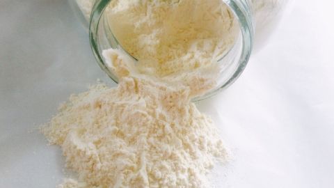 Ein Glas mit glutenfreiem Mehl kippt, während das Mehl auf eine weiße Oberfläche rieselt, ideal für glutenfreies Backen.
