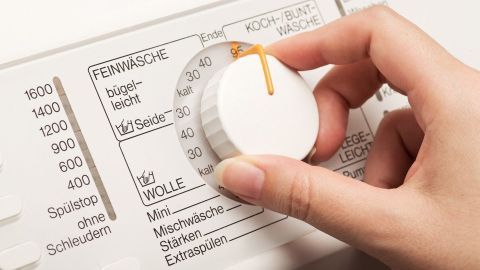 Eine Hand dreht den Waschmaschinenregler, um die Wäsche richtig zu waschen. Die Maschine steht in einem Wohnraum.