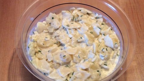 Ein klarer Glasschüssel mit cremigem Kartoffelsalat steht auf einem hölzernen Tisch, bereit zum Verzehr oder Aufpeppen.