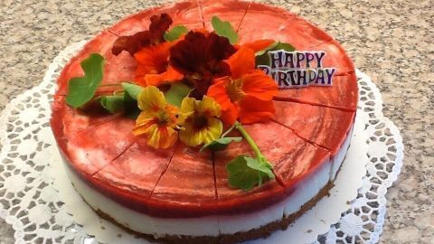 Eine Joghurt-Erdbeer-Torte steht dekoriert auf einem Teller, umgeben von bunten Blumen, mit der Aufschrift „Happy Birthday“.