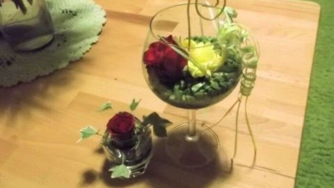 Ein großes Weinglas mit bunten Blumenarrangements steht auf einem Holz Tisch, umgeben von frischem Grün.