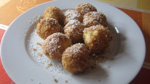 Runde Topfenknödel liegen auf einem weißen Teller, bestäubt mit Puderzucker, auf einem bunten Tisch.
