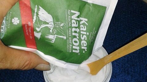 Eine Hand hält eine Tüte mit Kaiser-Natron über einen Behälter, während ein Holzstäbchen darin rührt, um Deo selbst zu machen.