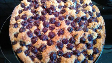 Ein runder Sauerkirschkuchen mit einer goldbraunen, krümeligen Oberfläche steht auf einer dunklen Oberfläche.