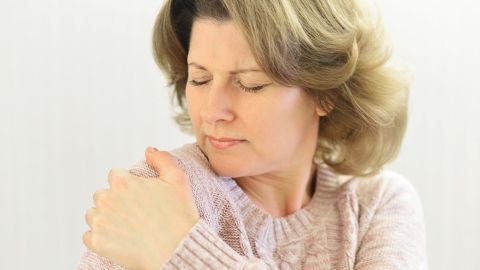 Eine Frau greift schmerzerfüllt an ihre Schulter, was auf eine Frozen Shoulder hindeutet, in einem hellen Raum.