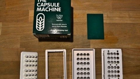 Eine Kapselmaschine steht auf einer Holzoberfläche, umgeben von Kapselhälften zur Befüllung mit Inhalt.