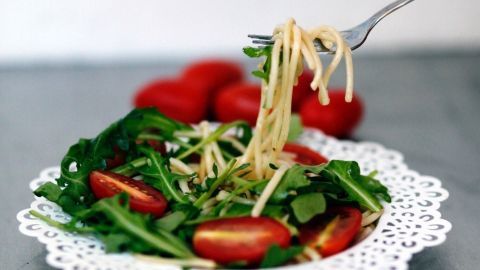 Eine Gabel hebt Pasta mit Rucola und Tomaten von einem dekorativen Teller, umgeben von frischen Zutaten in einem hellen Raum.