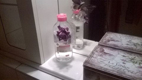 Eine Flasche Mizellen Reinigungswasser steht auf einem weißen Waschbecken neben einem dekorativen Behälter und einem Blumenstrauß.