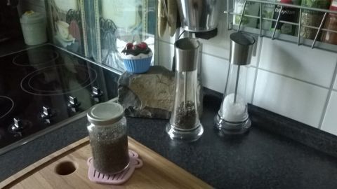 Ein Glas mit Chia-Samen steht auf einem Holzbrett in einer modernen Küche, umgeben von Gewürzmühlen und Kochutensilien.