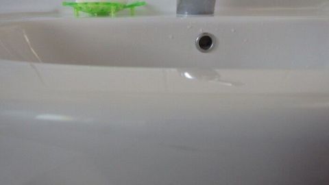 Ein sauberes Waschbecken mit Wasserstrahl, in einem hellen Badezimmer, während ein grünes Reinigungsscheuermittel daneben liegt.
