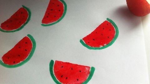Rote und grüne Kartoffeldrucke in Form von Wassermelonenstücken sind auf weißem Papier angeordnet, daneben liegt eine Kartoffel.