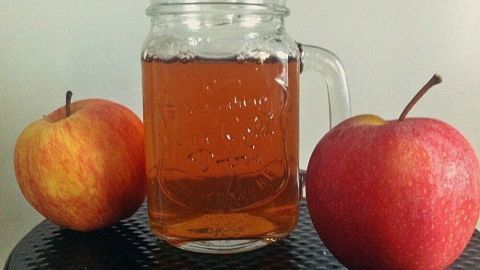 Ein Glas Apfelwein steht zwischen zwei frischen Äpfeln auf einem Tisch, umgeben von neutralem Hintergrund.