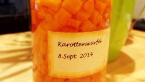 Ein Glas mit gewürfelten Karotten steht auf einem Holzuntergrund, umgeben von Pflanzen. Auf dem Etikett steht "Karottenwürfel 8. Sept. 2014".
