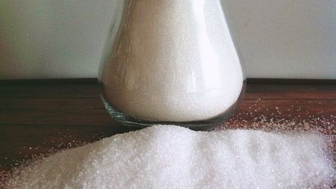 Ein Zuckergefäß steht auf einem Holztisch und hat einen Löffel, während der Zucker sich daneben verstreut.