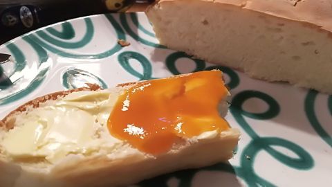 Ein auf einem Teller liegendes Stück Hefeteigstriezel mit Butter und Marmelade, ergänzt durch frisches Brot im Hintergrund.