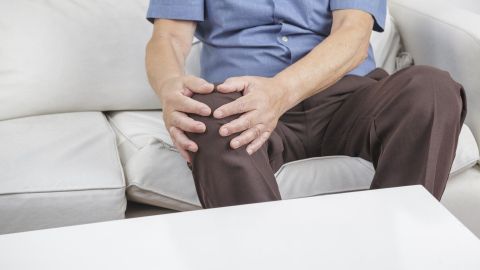 Ein Mann sitzt auf einem Sofa und hält sich schmerzhaft das Knie, während er sich offensichtlich unwohl fühlt.