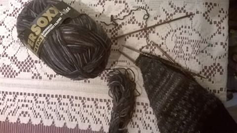 Ein Wollknäuel liegt auf einer Spitzendecke, während Stricknadeln ein begonnenes Projekt in Schwarz voranbringen.