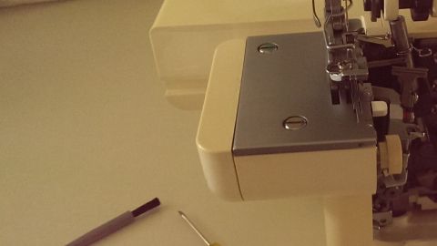 Eine Overlockmaschine steht bereit, während eine Person Nadeln wechselt. Werkzeuge wie Schraubendreher und Nadeln liegen daneben.