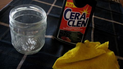 Etiketten-Kleberreste entfernen mit Glaskeramikreiniger. Ein leeres Glas steht neben einer Flasche mit CeraClen und einem gelben Tuch, ideal zum Entfernen von Etiketten-Kleberresten.