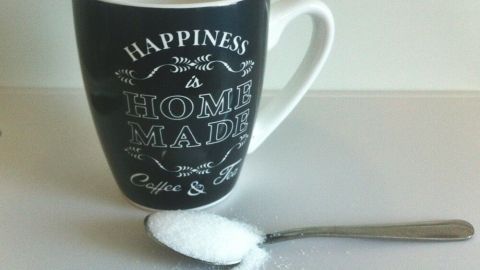 Eine schwarze Tasse steht auf einem Tisch, während ein Löffel mit Zucker daneben liegt, bereit zum Umrühren.