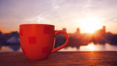Eine dampfende rote Tasse steht in der Morgensonne auf einem Holztisch, umgeben von einer städtischen Skyline.