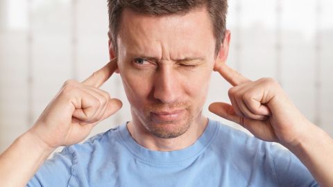 Ein Mann hält sich die Ohren zu, während er eine schmerzhafte Miene macht, als würde er unter Tinnitus leiden.
