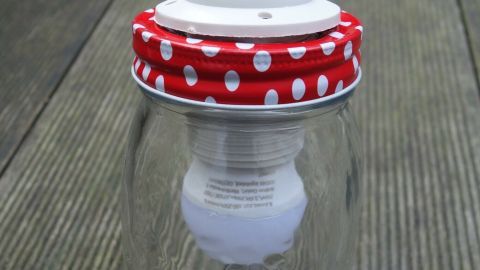 Eine Lampe aus einem Marmeladenglas hängt an einem Kabel und steht auf einem hellen Holzuntergrund.