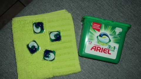 Vier Ariel 3in1 Pods liegen auf einem grünen Tuch, neben einer Verpackung in einem neutralen Umfeld.