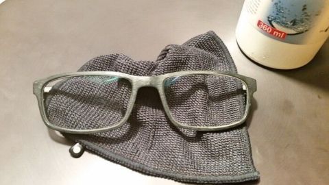Eine Brille liegt auf einem dunklen Reinigungstuch, während im Hintergrund eine Flasche mit Kontaktlinsen Pflegemittel steht.