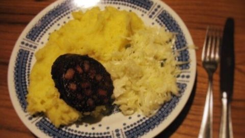 Blutwurst liegt appetitlich auf einem Teller, umgeben von cremigem Kartoffelpüree und frischem Krautsalat, auf einem Holzstuhl.