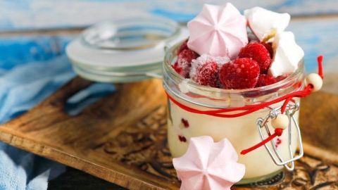 Ein Glas Himbeerdessert steht auf einem Holztablett, gefüllt mit frischen Himbeeren und bunten Baisers, vor einem blauen Hintergrund.