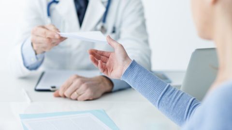 Ein Arzt übergibt einem Patienten ein Rezept in einem hellen, modernen Sprechzimmer. Dokumente liegen auf dem Tisch.