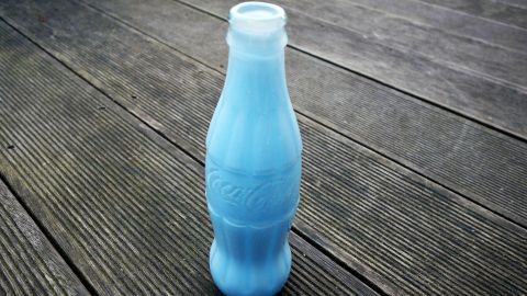 Eine bunte Colaflasche steht auf einem Holzfußboden und dient als Vase, kreativ umfunktioniert für Blumen.