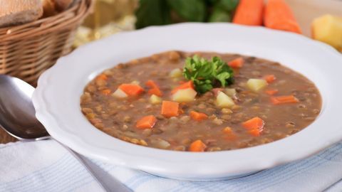 Eine Schüssel Linsensuppe mit braunen Tellerlinsen und buntem Gemüse steht auf einem Tisch, umgeben von Brot und frischem Gemüse.