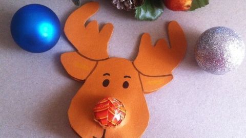 Ein süßes Rentier aus Papier mit einem Lolli als Nasenlicht, umgeben von Weihnachtskugeln auf einem festlichen Tisch.